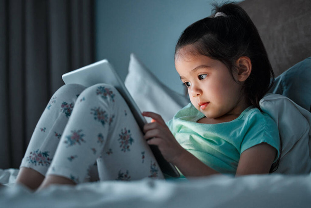 a little girl using a digital tablet while lying on her bed. - Foto, Bild