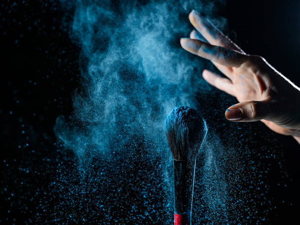Woman shaking off blue powder from a brush on a black background - Фото, зображення