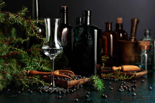 Gin borókamagokkal és ágakkal egy régi asztalon. A háttérben konyhai eszközök és antik üvegek vannak.. - Fotó, kép