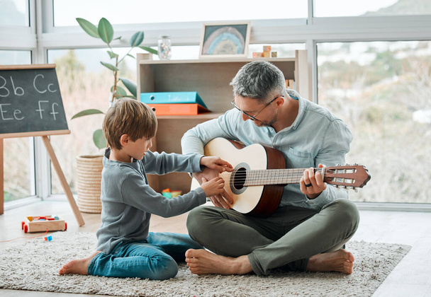 a young father teaching his son how to play the guitar while sitting on the floor at home. - Foto, Bild