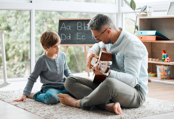 Ένας νεαρός πατέρας μαθαίνει στο γιο του πώς να παίζει κιθάρα ενώ κάθεται στο πάτωμα στο σπίτι.. - Φωτογραφία, εικόνα
