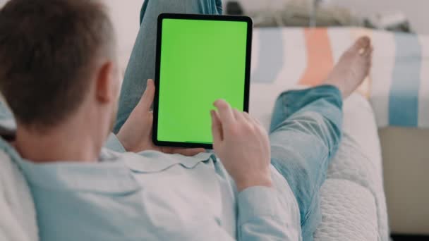 hombre irreconocible acostado en un sofá con camisa casual usando tableta digital con pantalla verde tiempo de ocio en casa - Metraje, vídeo