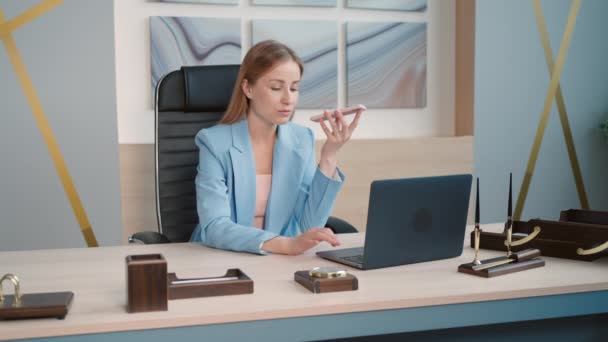 mujer de negocios exitosa sentada en un escritorio que sostiene el teléfono inteligente utilizando la ayuda de voz o enviar un mensaje en la oficina moderna - Imágenes, Vídeo