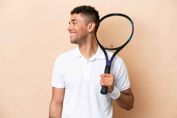 Young brazilian handsome man playing tennis isolated on beige background looking side - Фото, зображення