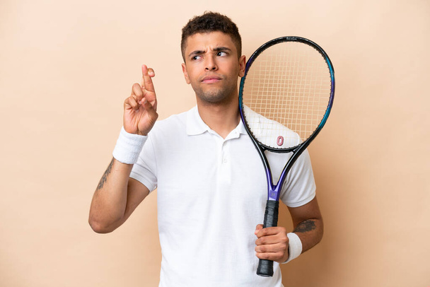 Young brazilian handsome man playing tennis isolated on beige background with fingers crossing and wishing the best - Photo, Image