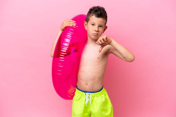 Petit garçon caucasien tenant un beignet gonflable isolé sur fond rose montrant pouce vers le bas avec expression négative - Photo, image