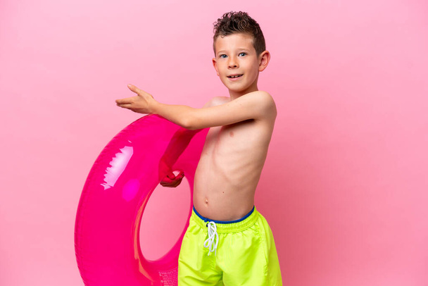 Pequeño chico caucásico sosteniendo una rosquilla inflable aislada sobre fondo rosa extendiendo las manos a un lado para invitar a venir - Foto, imagen