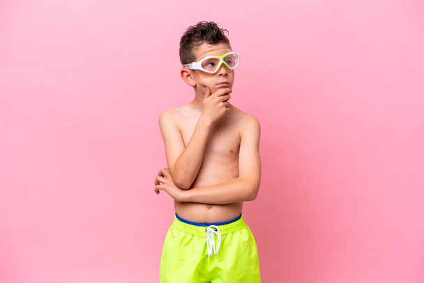 Little caucasian boy wearing a diving goggles isolated on pink background having doubts - Photo, Image