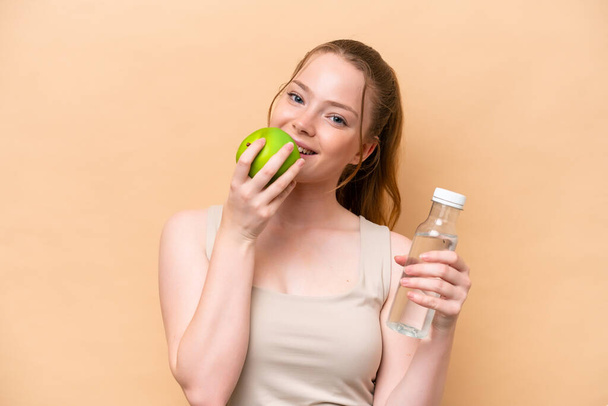 Young caucasian girl isolated on beige background with a bottle of water and eating an apple - Foto, imagen