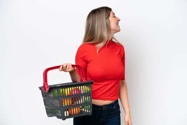 Young Rumanian woman holding a shopping basket full of food isolated on white background laughing in lateral position - Фото, изображение