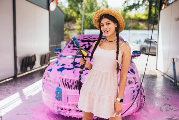 Молода жінка шланг високого тиску стоїть перед автомобілем, покритим рожевою піною
 - Фото, зображення
