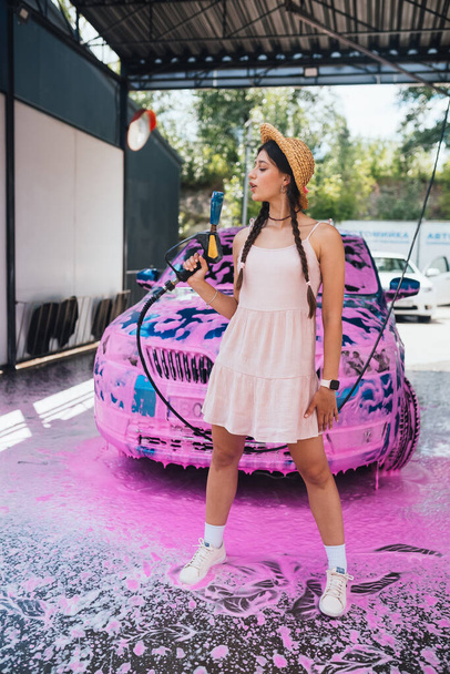 Young woman high pressure hose stands in front of a car covered in pink foam - Foto, afbeelding