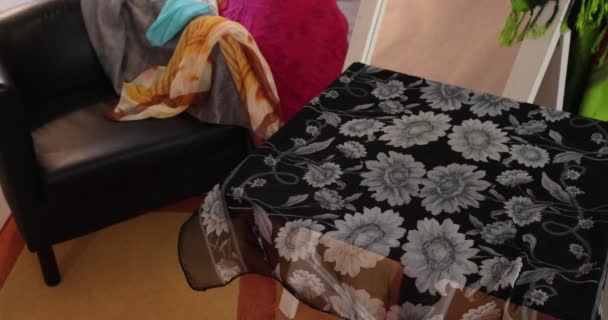Hair straightening equipment placed by a woman's hand on a small table covered with a floral scarf, straightener iron, brush and comb, room with a bench, a mirror and scattered multi-colored scarves - Metraje, vídeo