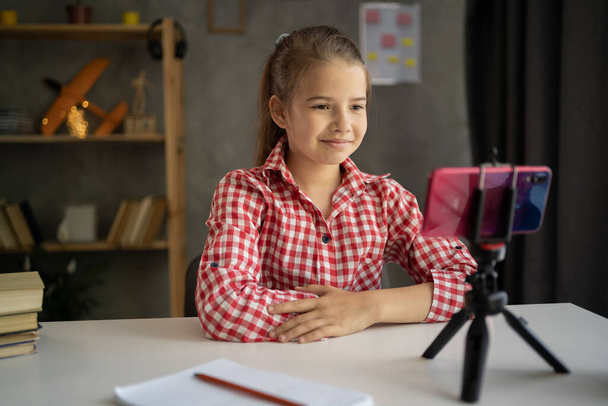 girl student studying online at home, looking at phone screen, recording video talking to cellphone on tripod, watching webinar, using smartphone on tripod, writing notes, homeschooling concept - Photo, Image