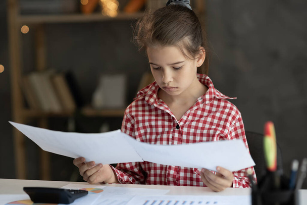 Focused young girl child looking through paper documents, managing business affairs, summarizing taxes, planning future investments, accounting alone at home office. Achievement career concept - Фото, изображение