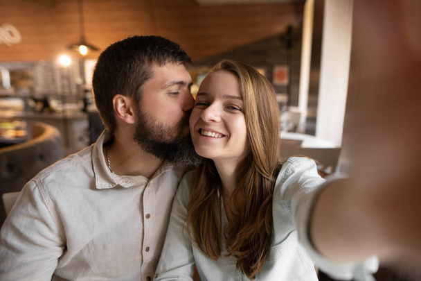 Smiling beautiful woman and her handsome boyfriend. Couple at their date in restaurant. Happy cheerful family. They taking Pov selfie at cafe and kissing - Fotografie, Obrázek