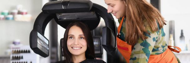 Portrait of young lady under hairdressing machine while looking in mirror. Visit master for beautiful look and good hair quality. Self care, beauty concept - Foto, Imagen