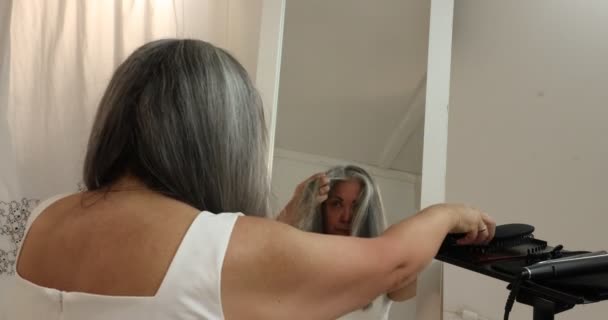 Зрелая женщина концентрируется на процессе выпрямления волнистых седых волос самостоятельно, скользя выпрямляющим железом по нитке, отраженной в зеркале на белом фоне - Кадры, видео
