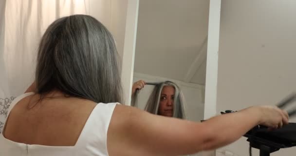 Mujer madura deslizando el hierro alisado sobre una hebra de su cabello, concentrándose en el proceso de alisar el cabello negro gris ondulado por sí misma, reflejado en el espejo sobre un fondo blanco - Imágenes, Vídeo