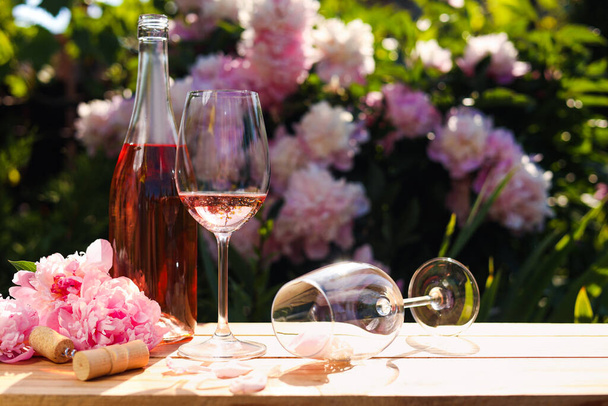 Bottle and glass of rose wine near beautiful peonies on wooden table in garden. Space for text - Foto, Imagem