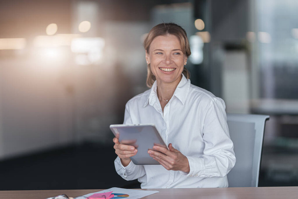 Gözlüklü gülümseyen iş kadını dijital tableti tutuyor ve modern ofise bakıyor. - Fotoğraf, Görsel