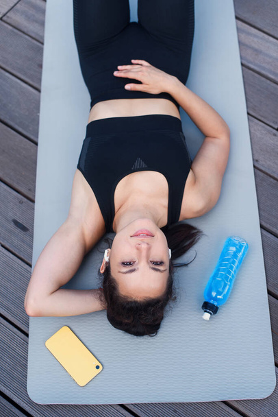 Woman lying with phone and bootle of water on yoga mat after exercising, relaxing with smartphone and listening to music. Workout Playlist. - Fotografie, Obrázek