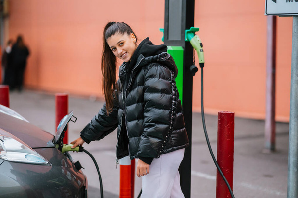 Młoda piękna kobieta podróżuje samochodem elektrycznym zatrzymując się na stacji ładowania stojąc podłączając kabel przeglądając internet na smartfonie uśmiechając się radosny podczas ładowania - Zdjęcie, obraz