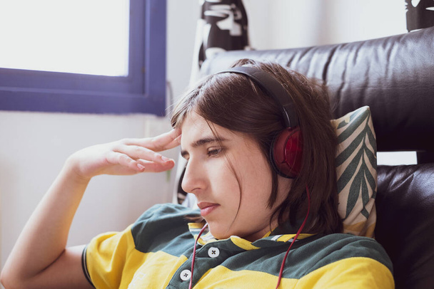 Грустный задумчивый подросток с длинными волосами, слушающий музыку в красных наушниках и склоняющийся головой к руке - Фото, изображение