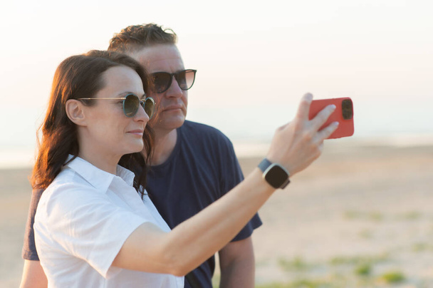 Yaş farkı olan mutlu bir çift. Genç bir kadın ve orta yaşlı bir adam sahilde selfie çekiyorlar. - Fotoğraf, Görsel