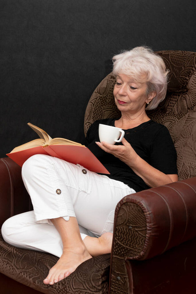 Een oudere mooie blonde vrouw van 60-65 jaar oud leest een boek, houdt een kopje in haar hand, zit in een fauteuil op een zwarte achtergrond. - Foto, afbeelding