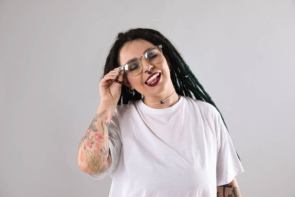 Beautiful young woman with tattoos on arms, nose piercing and dreadlocks against grey background - Zdjęcie, obraz