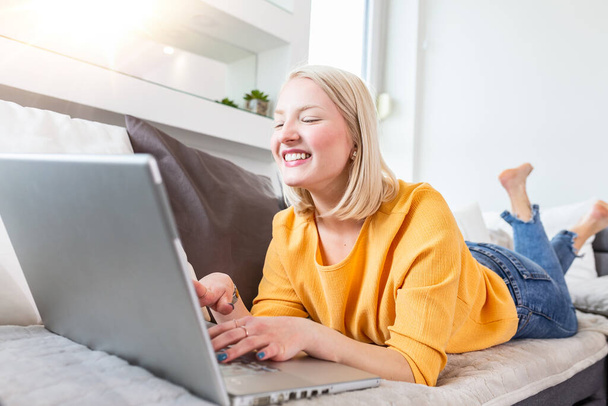 woman is relaxing on comfortable couch and using laptop at home.Portrait of beautiful young woman working on laptop while lying on sofa. Female using laptop at home. - Photo, Image