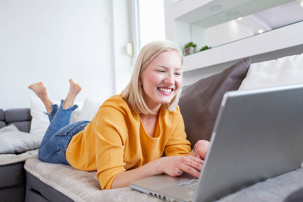 kobieta relaksuje się na wygodnej kanapie i korzystając z laptopa w domu.Portret pięknej młodej kobiety pracującej na laptopie leżącej na sofie. Kobieta korzystająca z laptopa w domu. - Zdjęcie, obraz