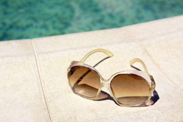 Stylish sunglasses near outdoor swimming pool on sunny day, closeup. Beach accessory - Fotó, kép