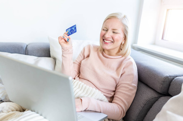 Nuori nainen sohvalla ostoksia verkossa pankkikortilla. Kaunis tyttö käyttää kannettavaa tietokonetta online-ostoksia kotona - Valokuva, kuva