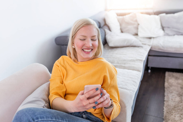 Полный снимок улыбающейся женщины, читающей сообщение на мобильном телефоне, сидящей дома на диване. Счастливая девушка читает сообщение в смартфоне, сидя дома на диване - Фото, изображение