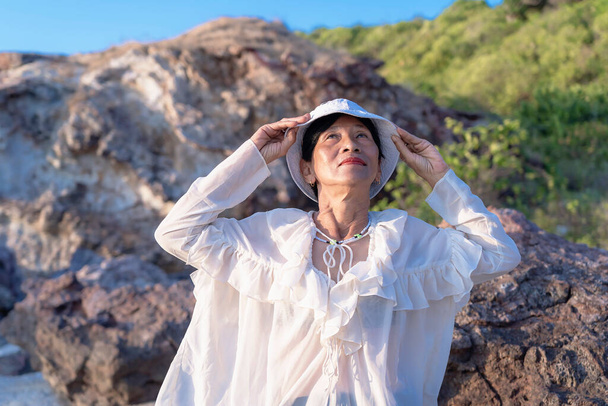 Portrait of a happy mature woman smiling outdoors. Happy smiling mature woman on the rock at sea beach, positive female wearing a white hat. Happiness, joy, relaxation, emotions, middle-aged people - Fotografie, Obrázek