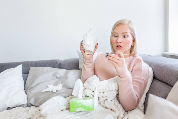 病気、季節のウイルス問題の概念。風邪をひいた女性は温度計で温度を見てソファに寝そべっている。病気の女性が高熱でベッドに横たわっている。風邪インフルエンザと片頭痛. - 写真・画像