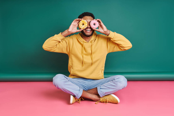 Playful man holding doughnuts in front of eyes while sitting against colorful background - Фото, изображение
