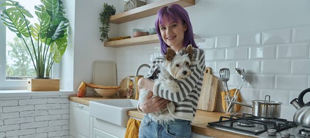 Cheerful young woman with purple hair carrying cute little dog while standing at the kitchen - Valokuva, kuva