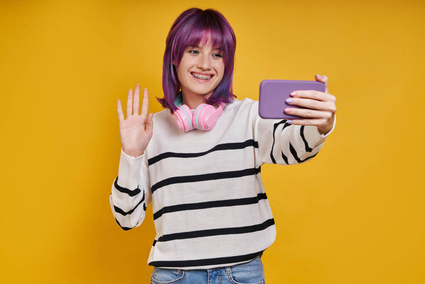 Cheerful young woman having video call while standing against yellow background - Φωτογραφία, εικόνα
