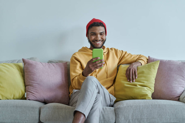 Handsome African man looking at his smart phone and smiling while sitting on the couch - Foto, imagen