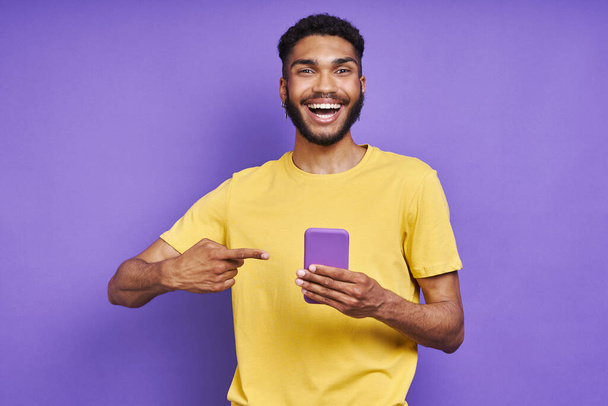 Happy African man pointing his smart phone while standing against purple background - Foto, imagen
