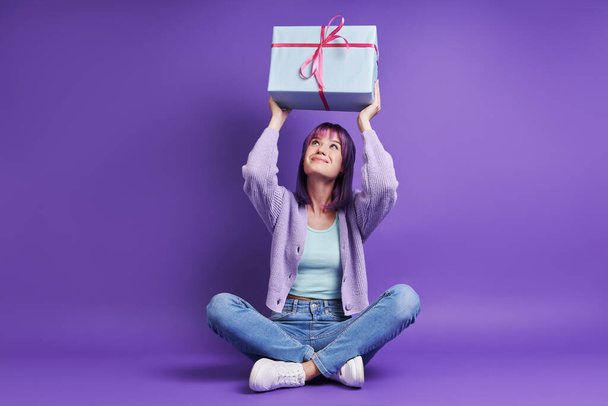 Beautiful young woman holding gift box upon head while sitting against purple background - Fotoğraf, Görsel