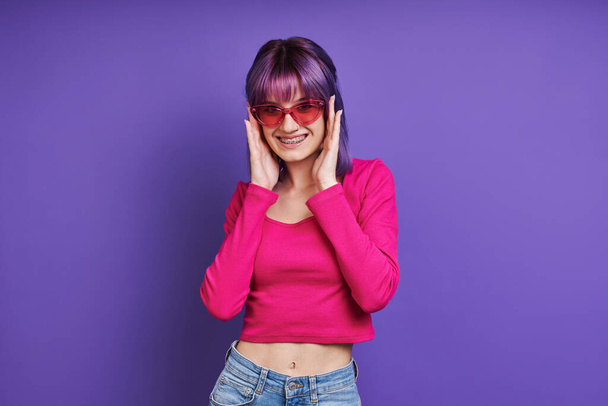 Cheerful young woman adjusting eyeglasses while standing against purple background - Foto, imagen