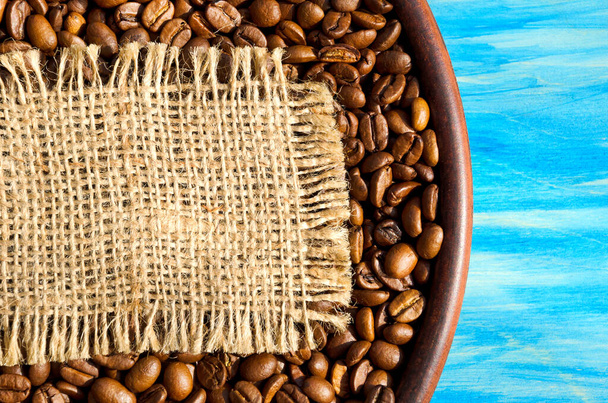 Roasted coffee beans in a ceramic plate on a turquoise background. A piece of burlap for text. Grains of a tropical plant. - Photo, image
