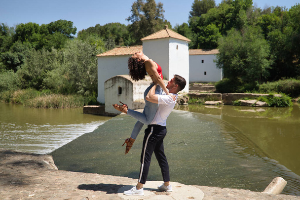 Ελκυστικό νεαρό ζευγάρι που χορεύει αισθησιακά μπάχατα σε ένα πέτρινο πάτωμα, σε ένα υπαίθριο πάρκο, δίπλα σε ένα ποτάμι. Λατινική έννοια χορού, αισθησιακό, λαογραφικό, αστικό. - Φωτογραφία, εικόνα