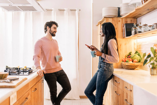 Beautiful black woman and her boyfriend using phone while standing in the kitchen at home. - Foto, Imagem