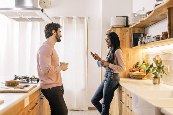 Beautiful black woman and her boyfriend using phone while standing in the kitchen at home. - 写真・画像