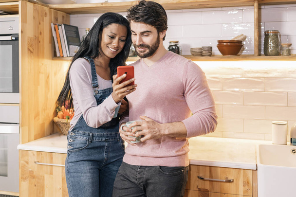 Beautiful black woman showing her boyfriend something in the phone while standing in the kitchen at home. - Foto, Imagem